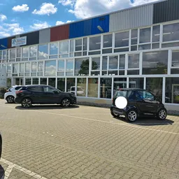 Parkeerplaats voor de gevel van kringloopwinkel met grote raampartijen en gekleurde dakstrepen