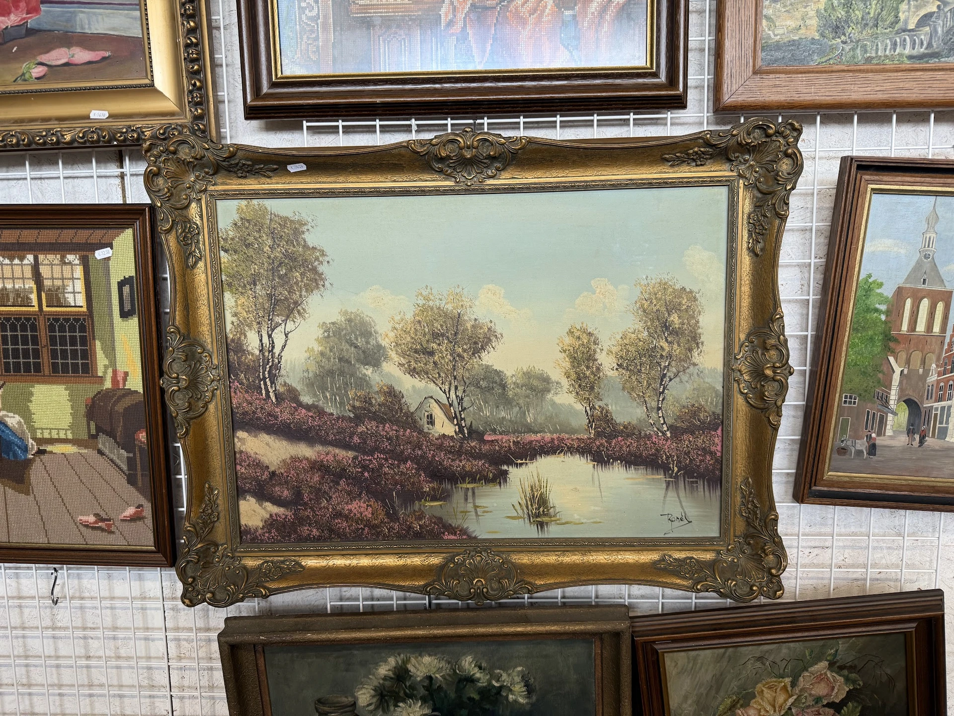 Ingelijste landschapsschildering met gouden barokke lijst, uitgestald tussen andere kunstwerken in een kringloopwinkel