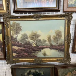 Ingelijste landschapsschildering met gouden barokke lijst, uitgestald tussen andere kunstwerken in een kringloopwinkel