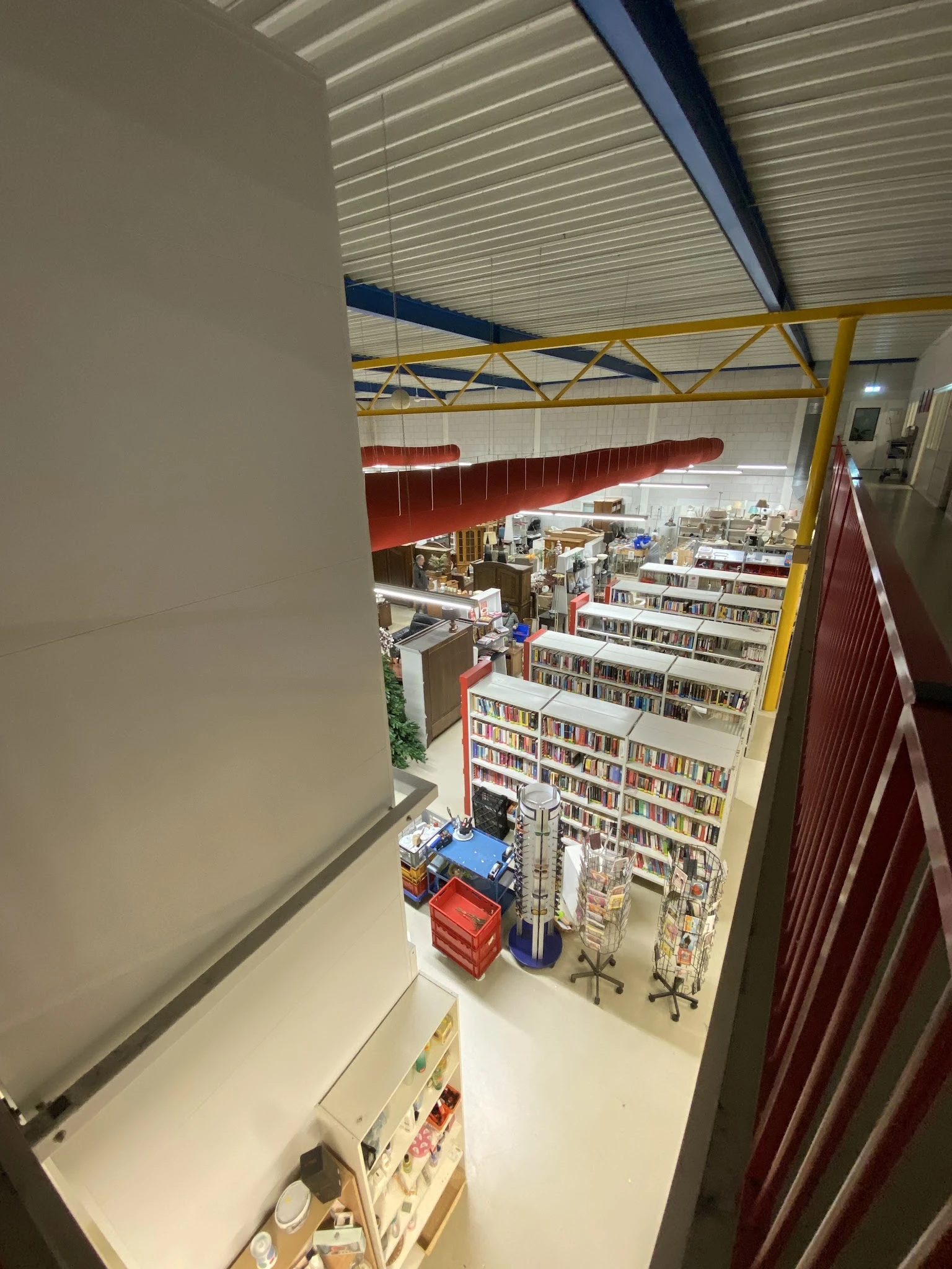 Overzicht van kringloopwinkel met boekenrekken, stellingen met diverse artikelen en geel geraamte aan plafond