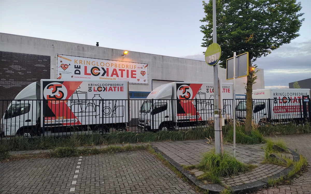 Voorgevel van kringloopwinkel met rode logo's en informatieborden achter een zwart hek, aan een straathoek met stoep en lanta