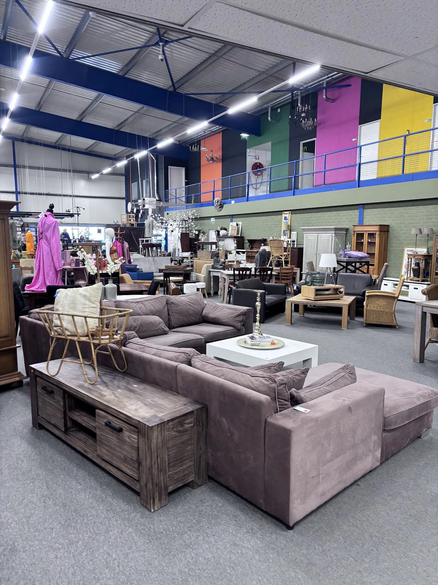 Ruim interieur van kringloopwinkel met grijze banken, houten tafels, stoelen en diverse meubels verspreid door de winkelruimt