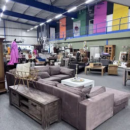 Ruim interieur van kringloopwinkel met grijze banken, houten tafels, stoelen en diverse meubels verspreid door de winkelruimt