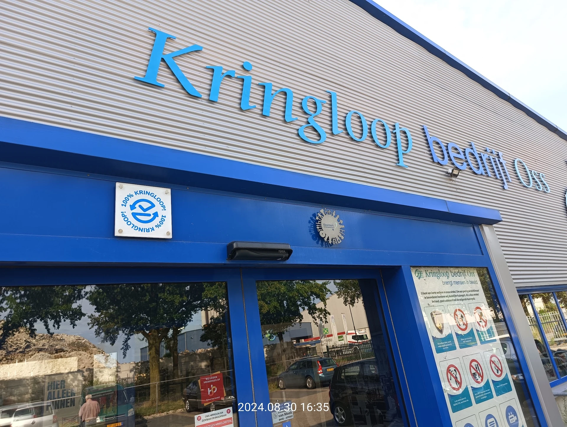 Blauwe gevel van kringloopwinkel met groot logo en ramen, straatgevel met planten