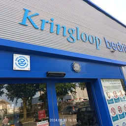 Blauwe gevel van kringloopwinkel met groot logo en ramen, straatgevel met planten