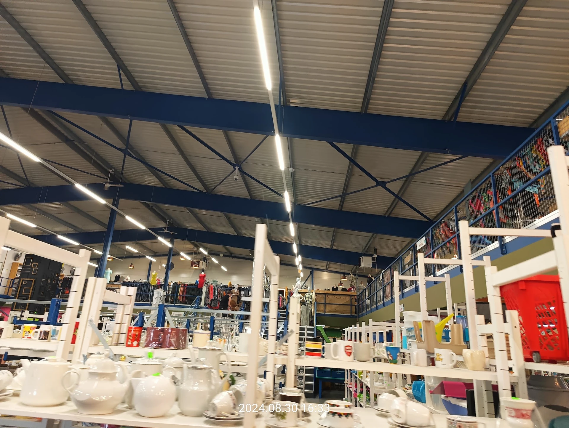 Ruim winkelinterieur met stellingen, witte serviesgoed in voorgrond, blauwe balken aan plafond, gekleurde bakken en spullen o