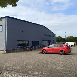 Voorgevel van kringloopwinkel met parkeerplaats en rode auto op de oprit