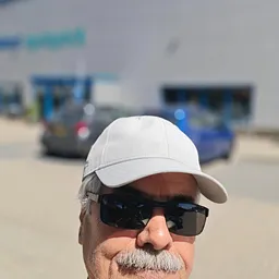 Selfie van een persoon met witte pet en zonnebril, op de achtergrond onscherpe voertuigen