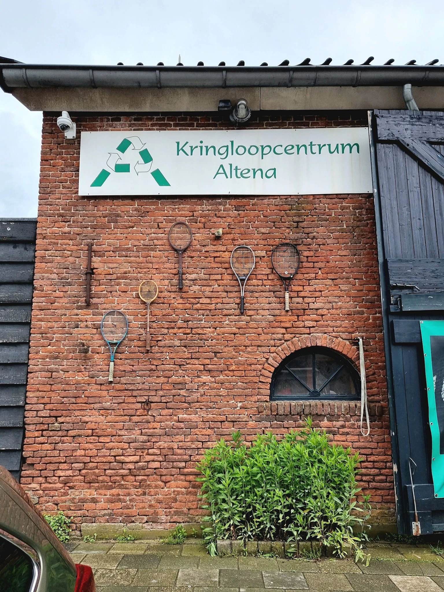 Bakstenen gevel met Kringloopcentrum Altena-logo en decoratieve tennisrackets aan de muur