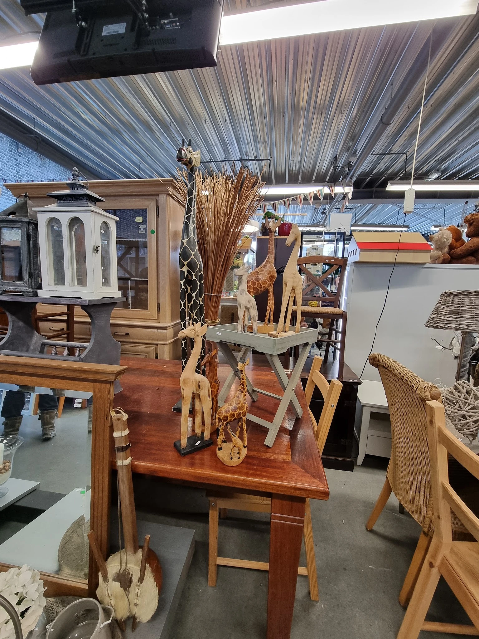 Houten decoratieve beeldjes en giraffen op een tafel in de kringloopwinkel, met droogbloemen en ander huisraad op de achtergr