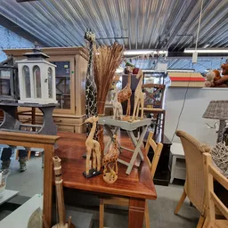 Houten decoratieve beeldjes en giraffen op een tafel in de kringloopwinkel, met droogbloemen en ander huisraad op de achtergr
