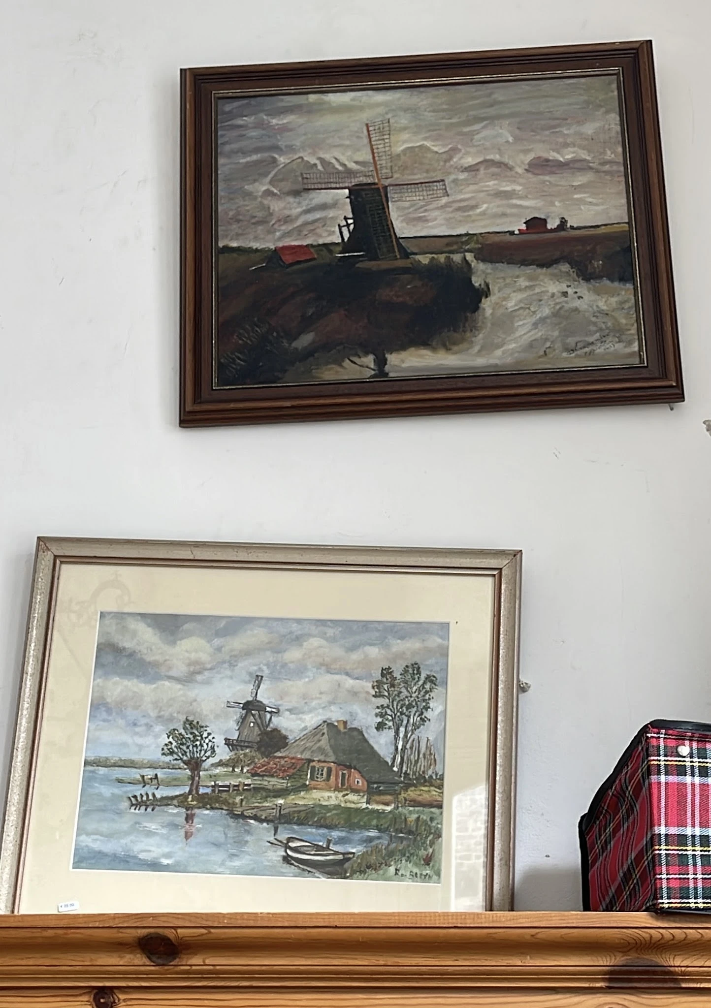 Twee ingelijste schilderijen aan een witte muur, waaronder afbeelding van een molen en landschap