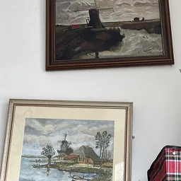 Twee ingelijste schilderijen aan een witte muur, waaronder afbeelding van een molen en landschap