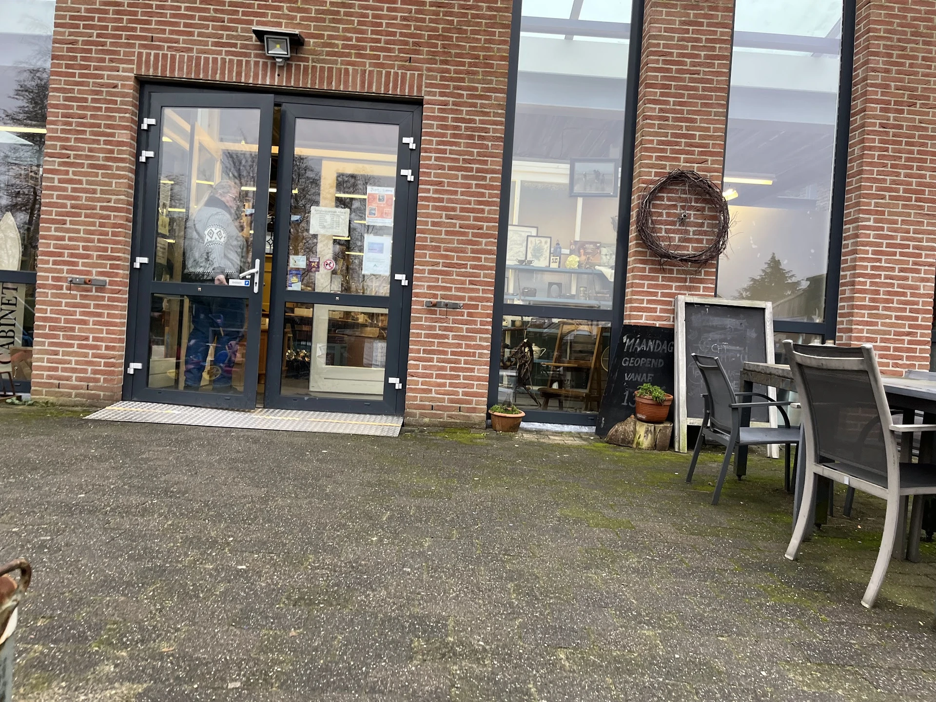 Bakstenen gevel van kringloopwinkel met zwarte deur, ramen en terras met stoelen