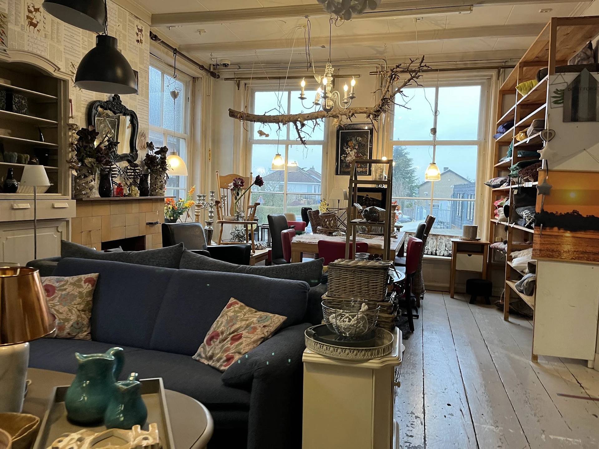 Ruim interieur van kringloopwinkel met zwarte bank, boekenkasten, hangende lampen en grote ramen met uitzicht