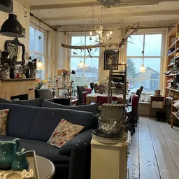 Ruim interieur van kringloopwinkel met zwarte bank, boekenkasten, hangende lampen en grote ramen met uitzicht