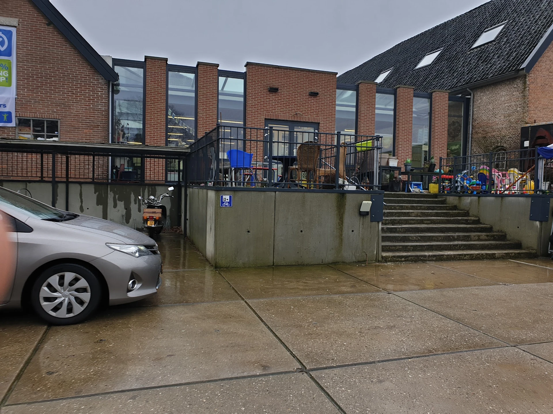 Voorkant kringloopwinkel met parkeerplaats, trap en ingang zichtbaar