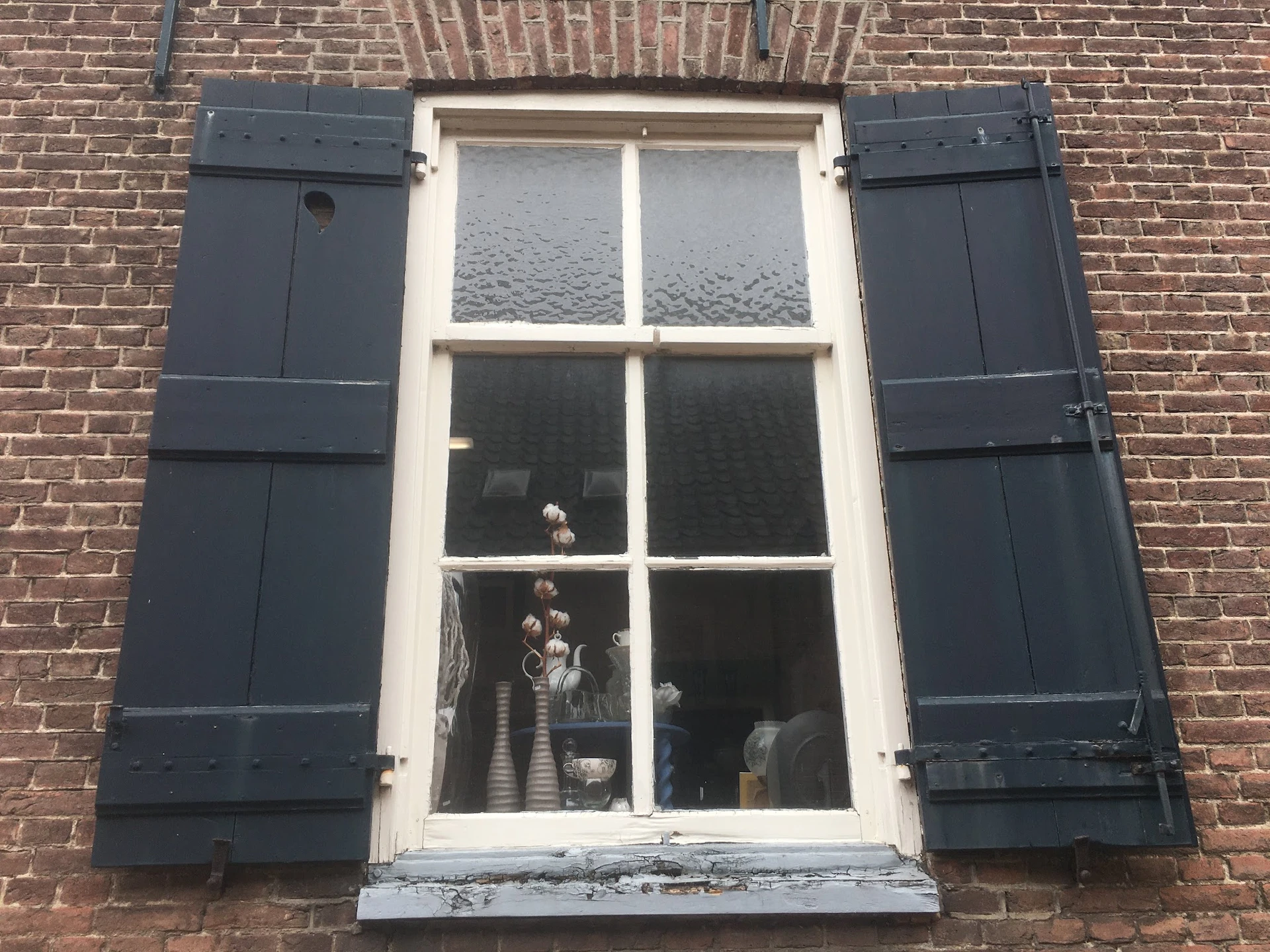 Bakstenen gevel met wit omkaderd raam en donkerblauwe luiken, details van woninginterieur zichtbaar
