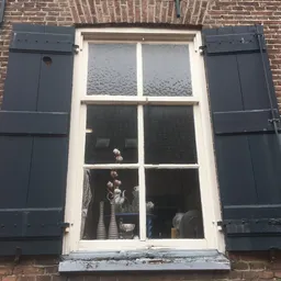 Bakstenen gevel met wit omkaderd raam en donkerblauwe luiken, details van woninginterieur zichtbaar