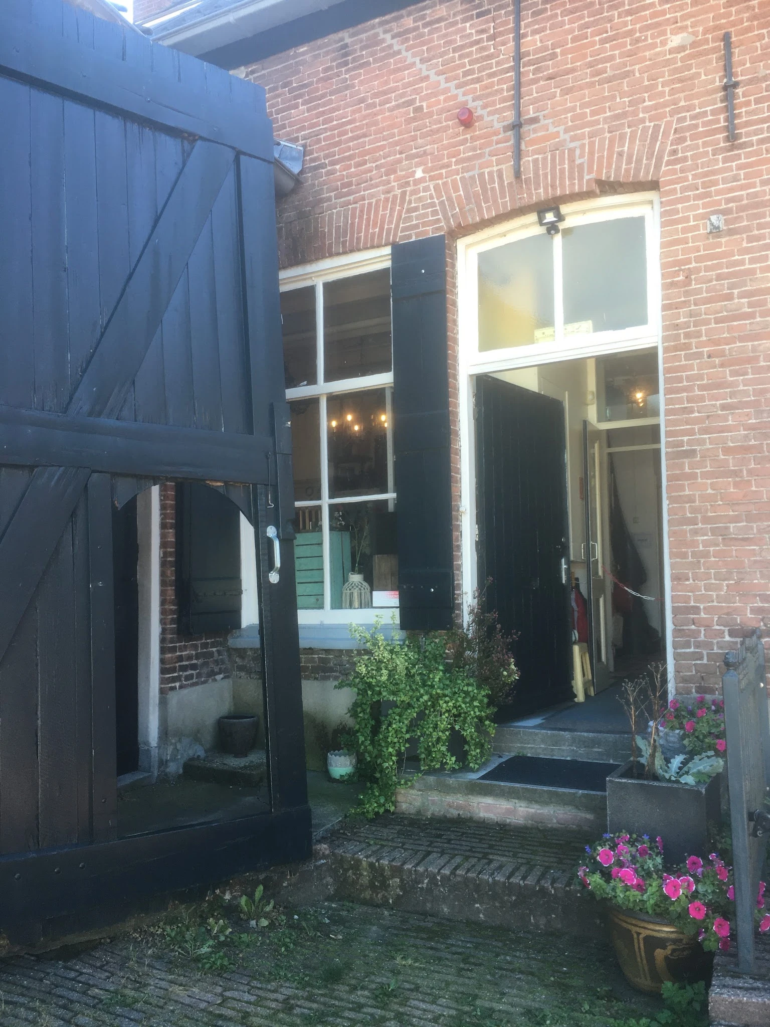 Ingang van een kringloopwinkel met zwarte deur en ramen in een bakstenen pand, met potplanten bij de ingang