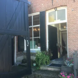 Ingang van een kringloopwinkel met zwarte deur en ramen in een bakstenen pand, met potplanten bij de ingang