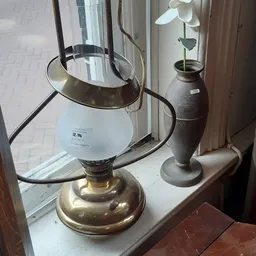 Twee vintage lampen op een vensterbank: een messing tafellamp met wit glazen kap en een donkere vaasachtige lamp met plantje