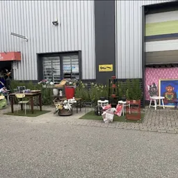 Voorkant van kringloopwinkel met stellingen en spullen buiten, roze muur en industrieel gebouw
