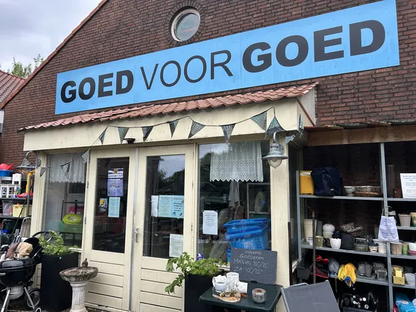 Gevel van kringloopwinkel met blauwe naam 'GOED VOOR GOED' boven de ingang, ramen met spullen zichtbaar