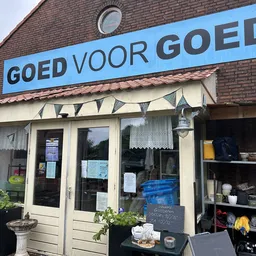 Gevel van kringloopwinkel met blauwe naam 'GOED VOOR GOED' boven de ingang, ramen met spullen zichtbaar