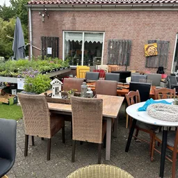 Terras met tweedehands meubilair uitgestald: tafels, stoelen en diverse meubels op het grasveld achter de winkel