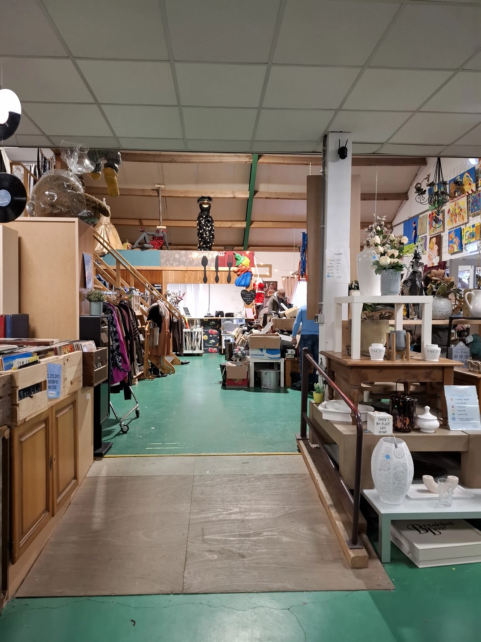 Winkelvloer van kringloopcentrum met groene vloer, kledingrekken, kasten, decoratie en verschillende afdelingen zichtbaar