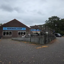 Bakstenen kringloopwinkel met blauwe KRINGLOOP-letters op gevel, aangrenzing met ander pand, geparkeerde auto, bewolkte hemel