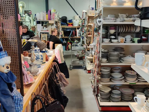 Kringloopwinkelinterieur met stellingen vol servies en borden rechts, toonbank links met klanten, diverse spullen en decorati