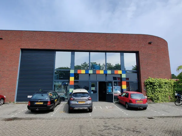 Voorkant van een bakstenen kringloopwinkel met grote ramen en gekleurde vierkanten, parkeerplaats met auto's