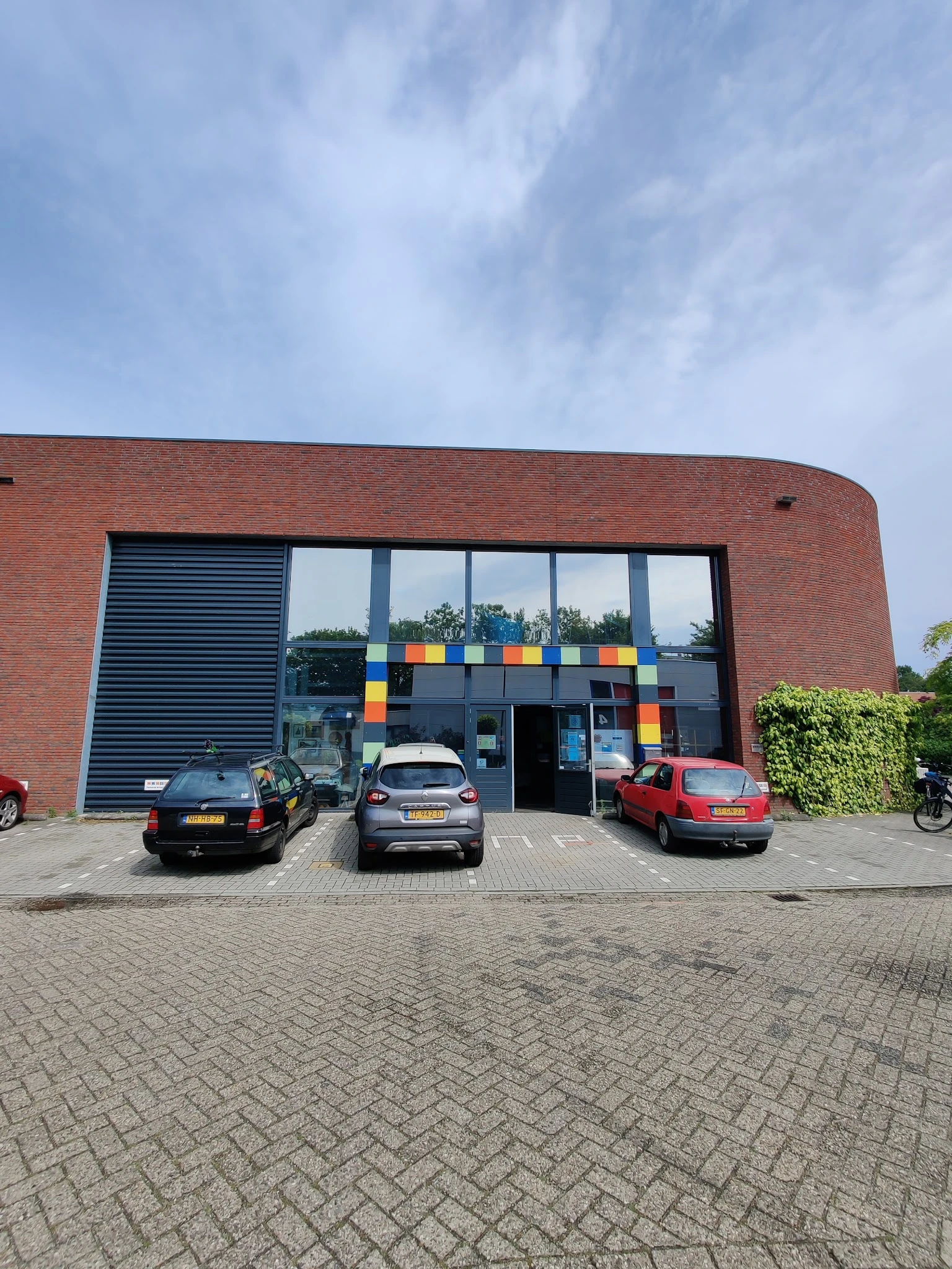 Voorkant van een bakstenen kringloopwinkel met grote ramen en gekleurde vierkanten, parkeerplaats met auto's