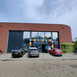 Voorkant van een bakstenen kringloopwinkel met grote ramen en gekleurde vierkanten, parkeerplaats met auto's