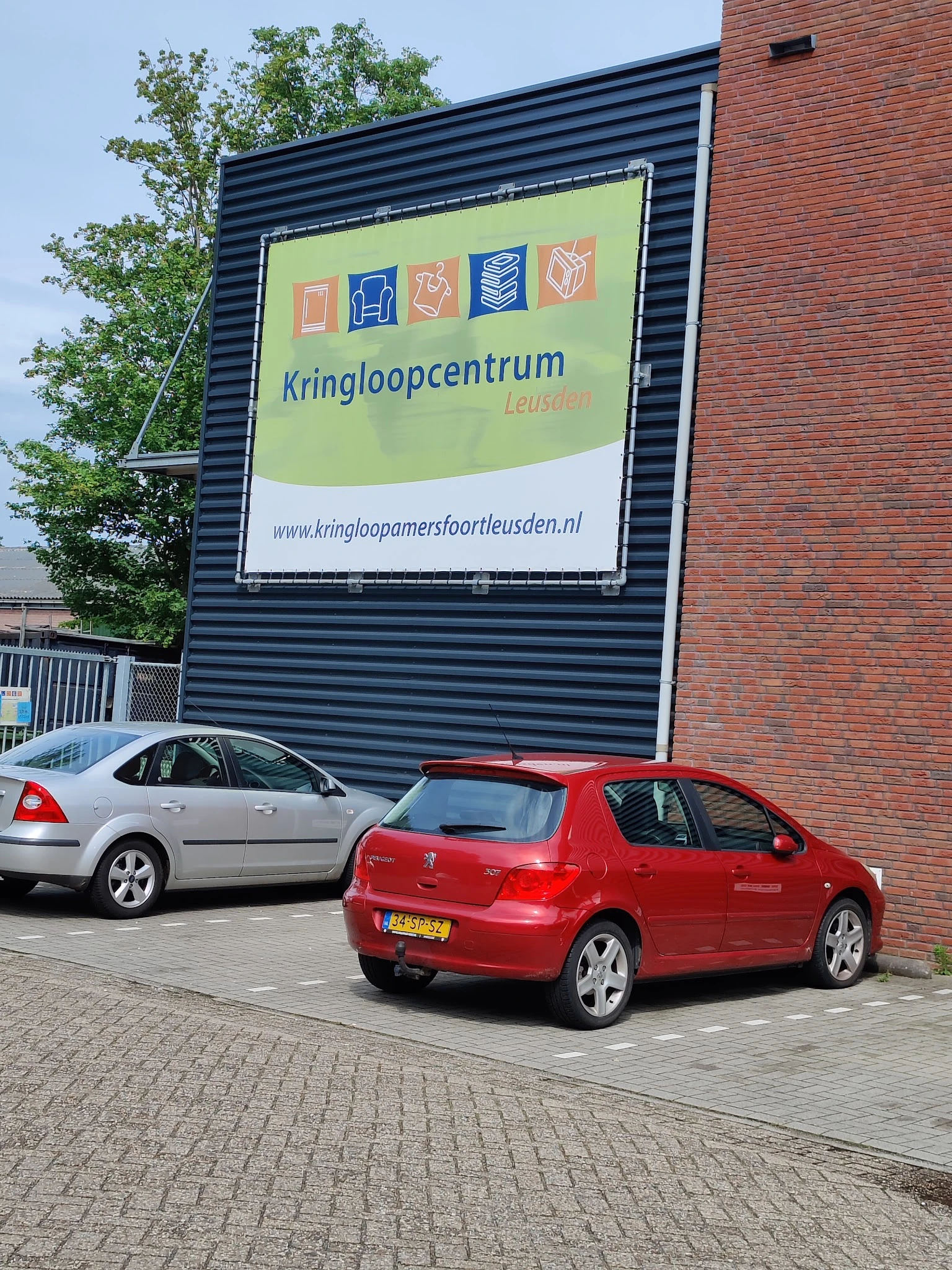 Voorzijde van kringloopwinkel met groen uithangbord, grijze golfplaten gevel en twee geparkeerde auto's