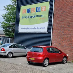 Voorzijde van kringloopwinkel met groen uithangbord, grijze golfplaten gevel en twee geparkeerde auto's