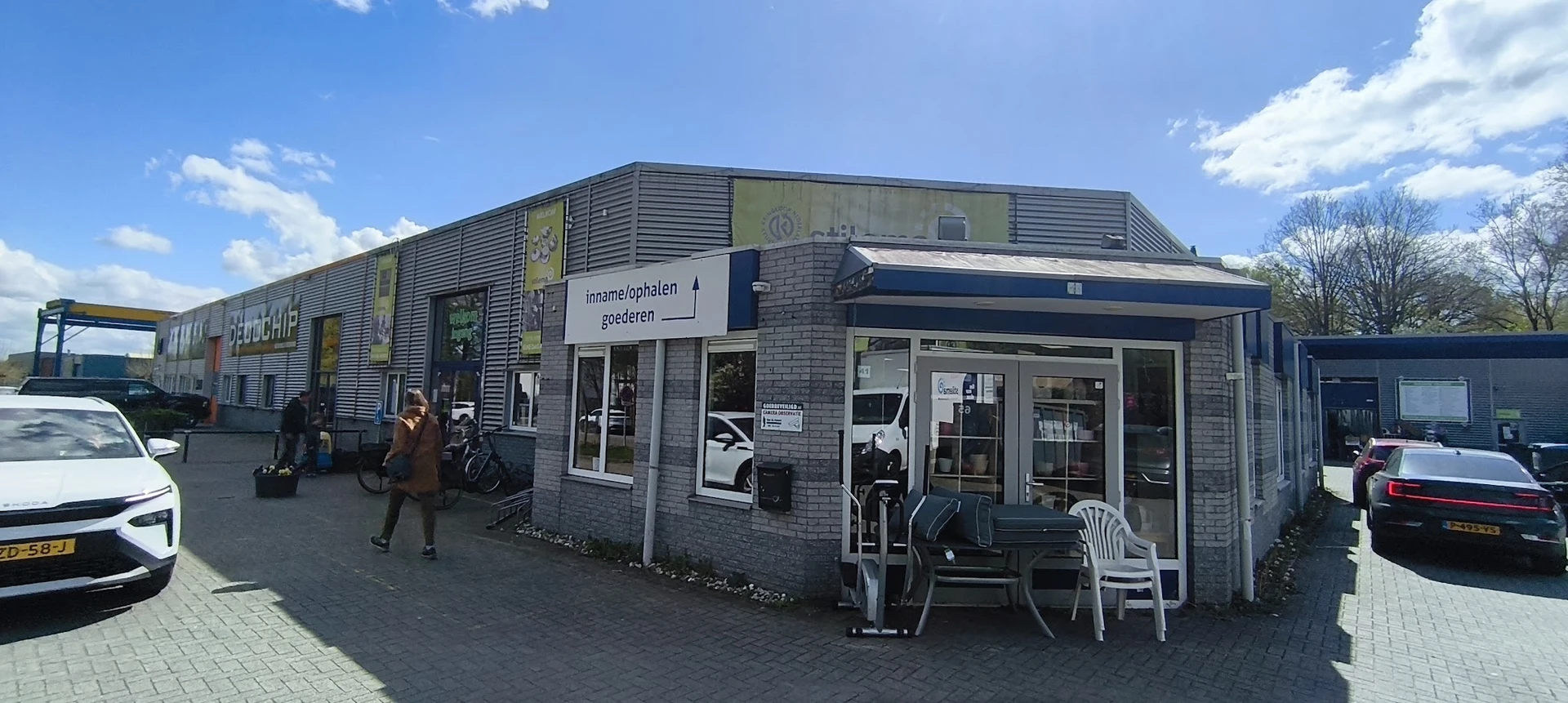 Voorgevel van kringloopwinkel met ingang, ramen en witte stoelen op het terras