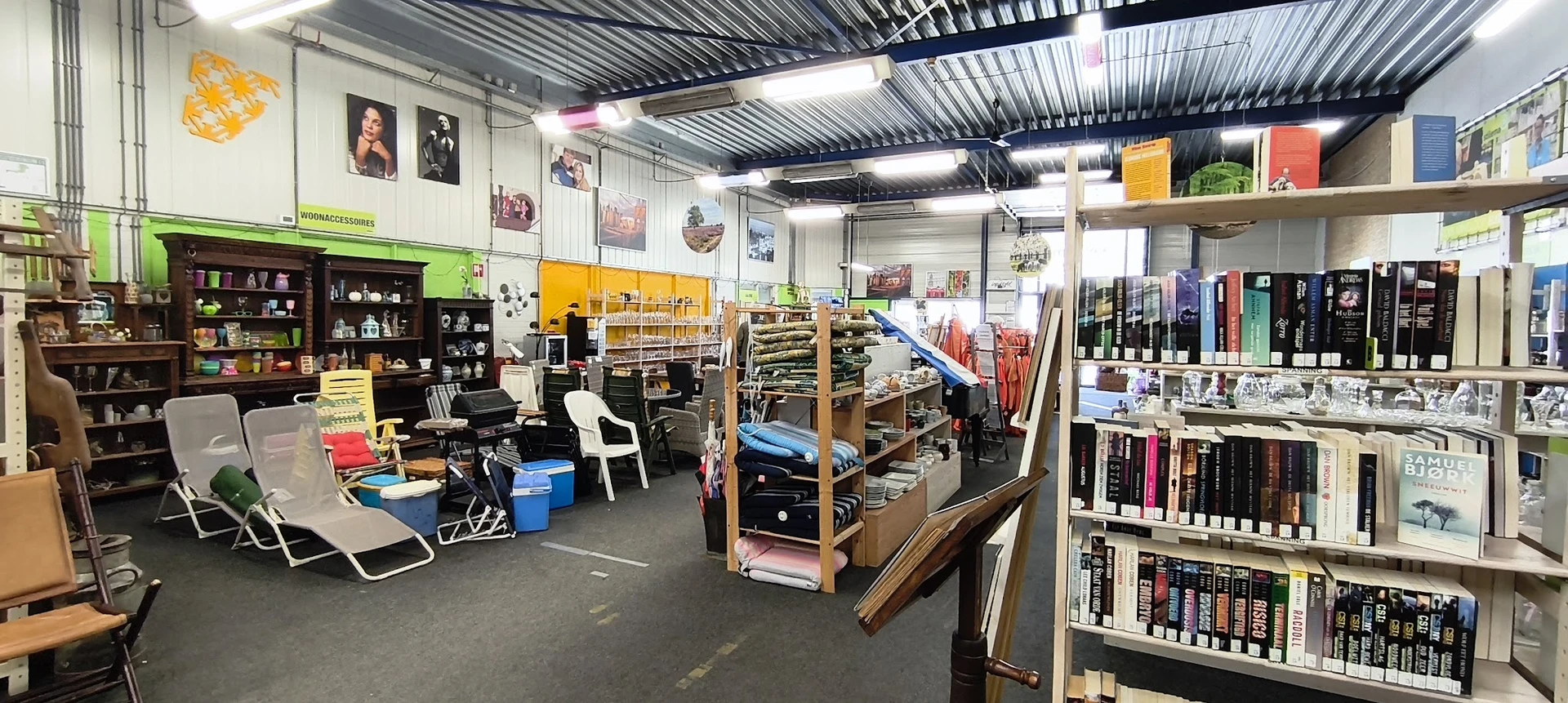 Ruim winkelbinnenin met stellingen vol spullen, kleding, boeken en diversen tegen de muren, grijze betonvloer en golfplaten p