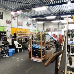 Ruim winkelbinnenin met stellingen vol spullen, kleding, boeken en diversen tegen de muren, grijze betonvloer en golfplaten p