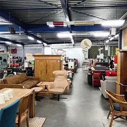 Winkelvloer met meubels: bruine leren bank, beige fauteuil, houten kasten en diverse zitmeubels op grijs beton