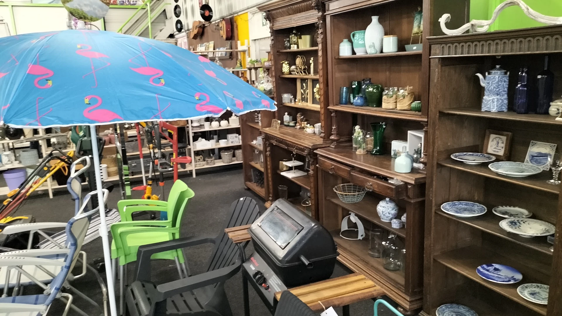 Winkelbinnenruimte met houten stellingkasten vol huishoudwaar, groene stoelen en blauwe parasol
