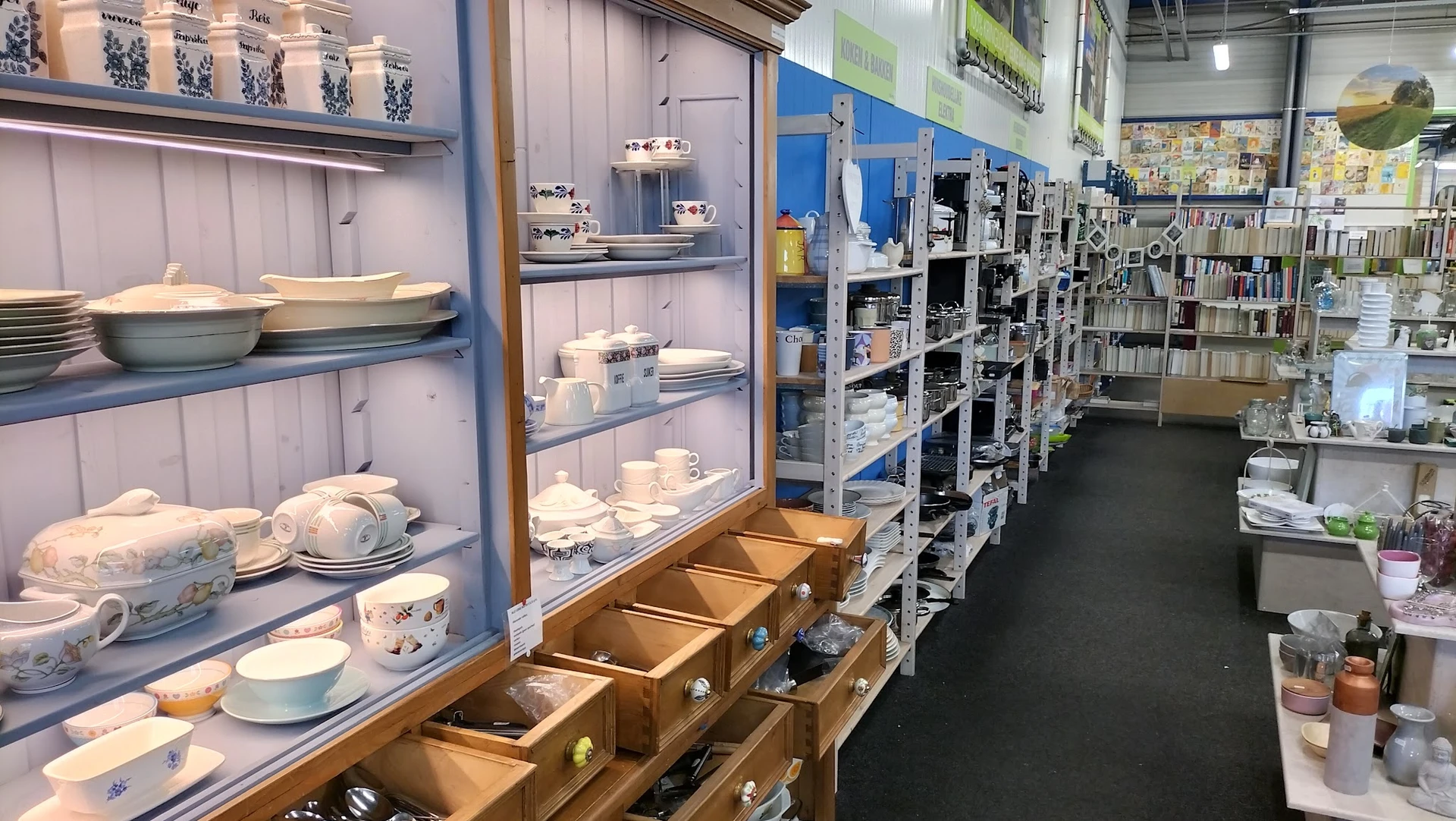 Vitrinekast met servies en kopjes, rechts stellingen met huishoudartikelen