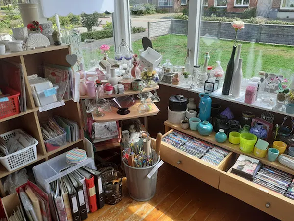 Hoek van kringloopwinkel met houten stellingen vol spullen, kasten met huishoudartikelen, bloemen en decoratie bij raam met u
