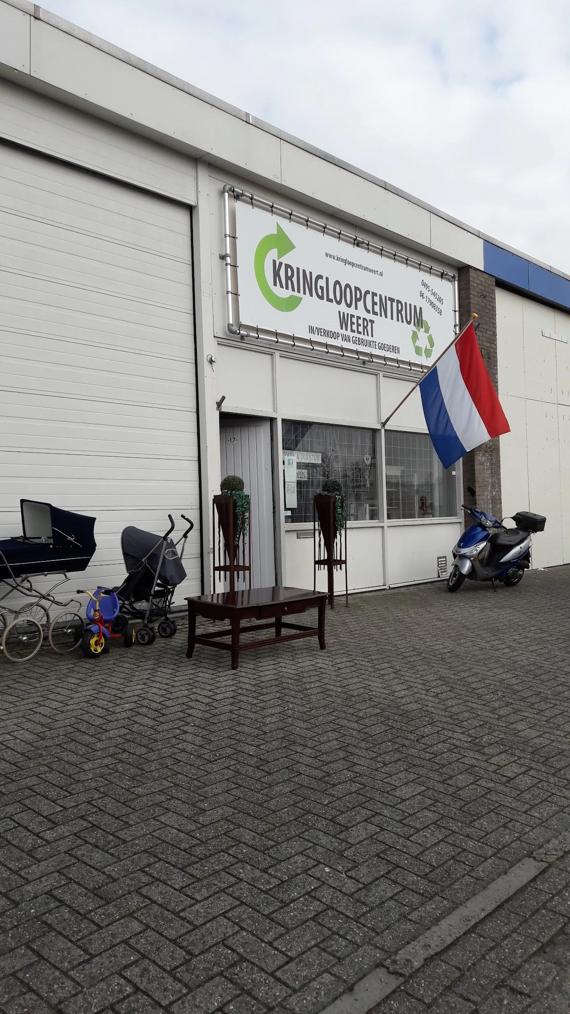 Voorgevel van kringloopwinkel met Nederlandse vlag, kinderwagen, fietsen en scooter op het terras