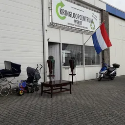 Voorgevel van kringloopwinkel met Nederlandse vlag, kinderwagen, fietsen en scooter op het terras