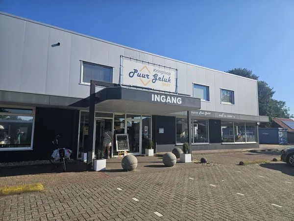 Gevel van kringloopwinkel met 'Pure Geluk' logo, ingang met zwarte deur en ramen, bakstenen plein ervoor