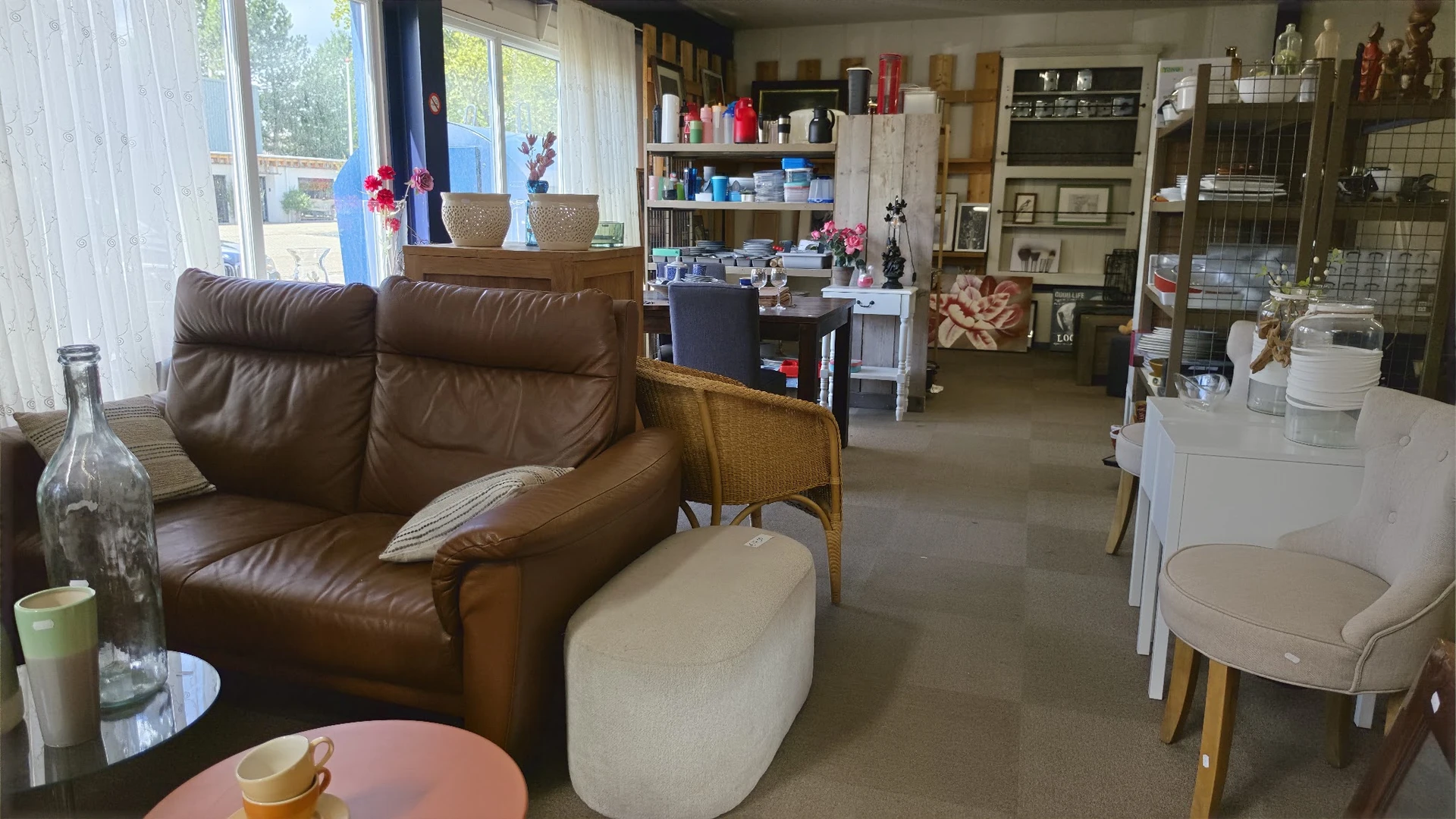 Winkelvloer met bruine leren fauteuil, witte poef, houten stoel, tafels, stellingen met diverse spullen en grote ramen met ui