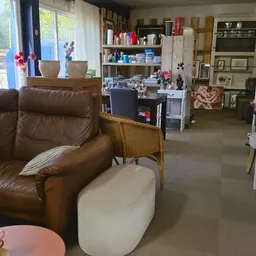 Winkelvloer met bruine leren fauteuil, witte poef, houten stoel, tafels, stellingen met diverse spullen en grote ramen met ui